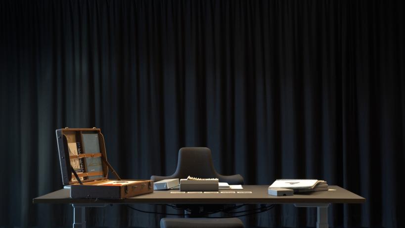 A desk stands in front of a dark curtain. Behind the table sits an empty chair, on the table lays an open briefcase, stacks of paper and different cards.