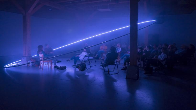 audience sat on chairs in a dimly lit room facing two speakers all illimunated by a ring light hanging from ceiling